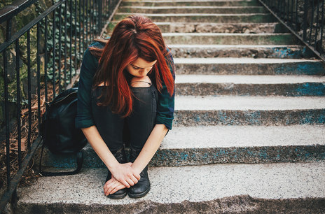 Eine Frau mit roten Haaren sitzt auf einer Treppe. Sie leht ihren Kopf traurig auf die Knie und schaut zum Boden.