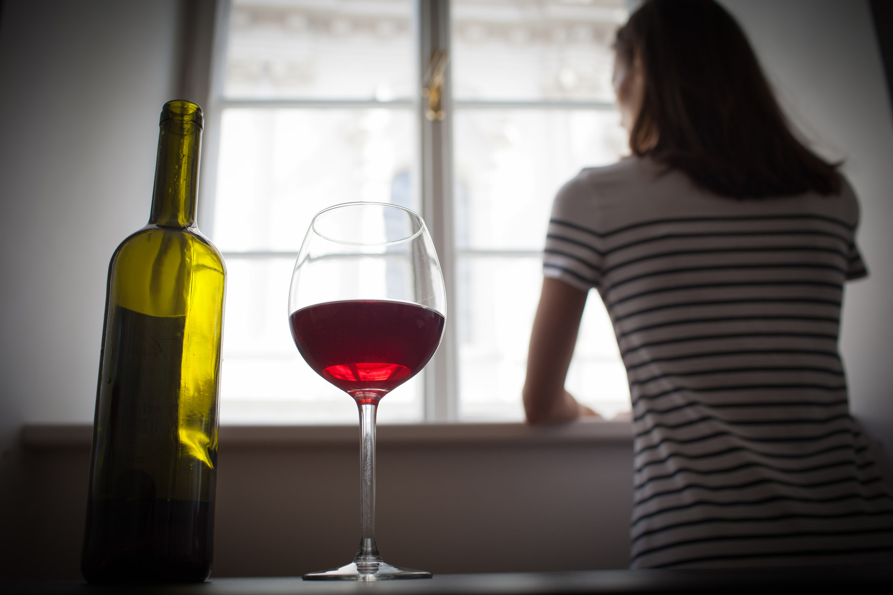 Im Vordergrund ein Weinglas und eine Flaschewein, im Hintergrund eine Frau, die aus dem Fenster schaut